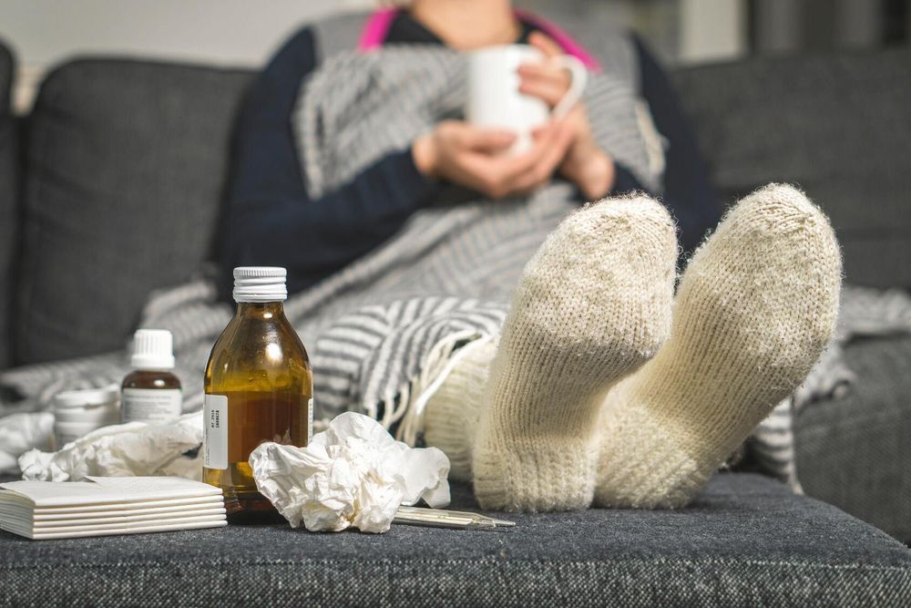 Person med filt och varma sockor, medicinflaskor bredvid.