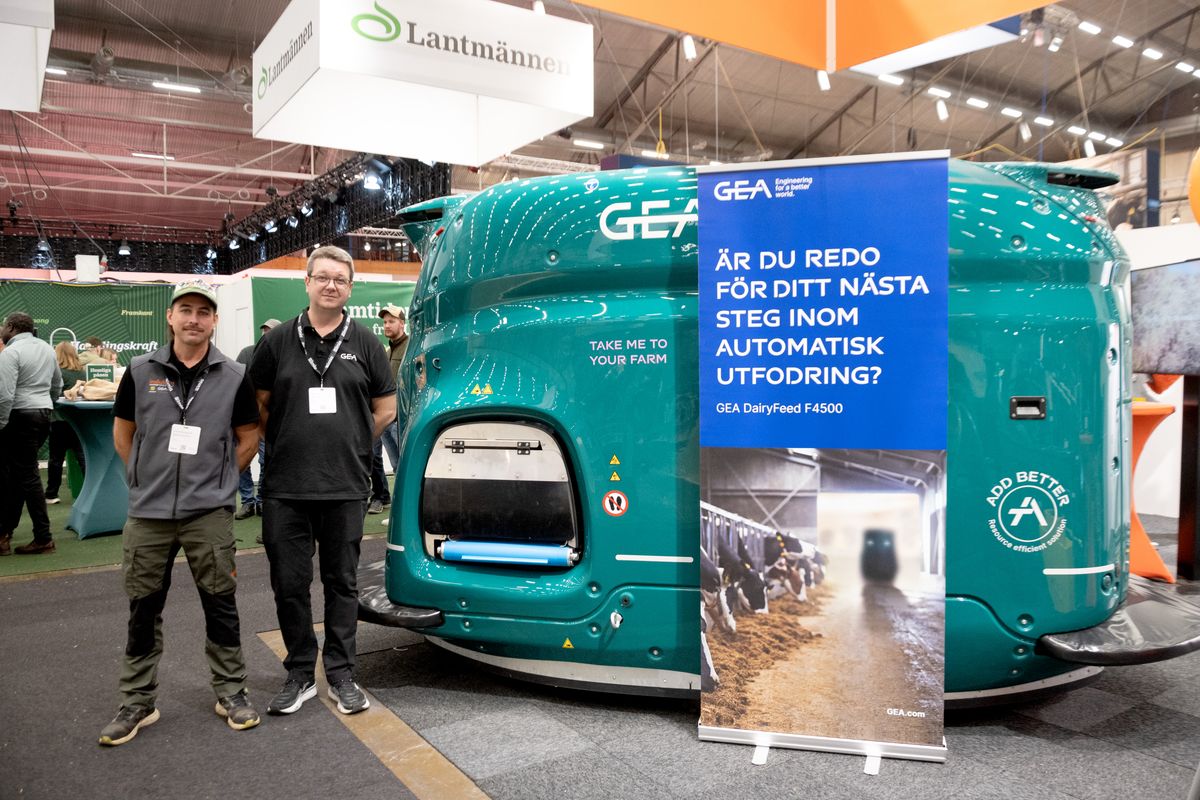 Två män står vid en grön automatisk utfodringsmaskin på en lantbruksmässa