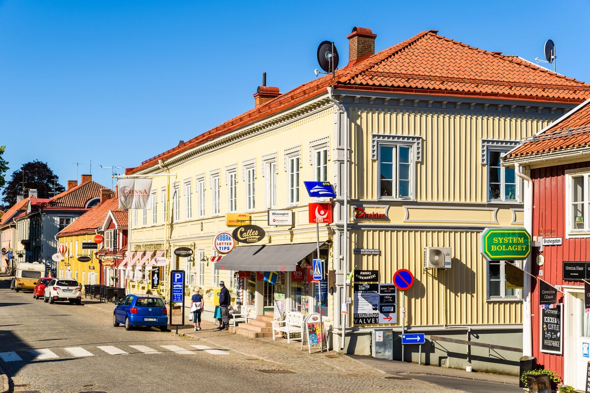 Småstadsgata med ljusa trähus, butiksfasader och gående längs trottoaren