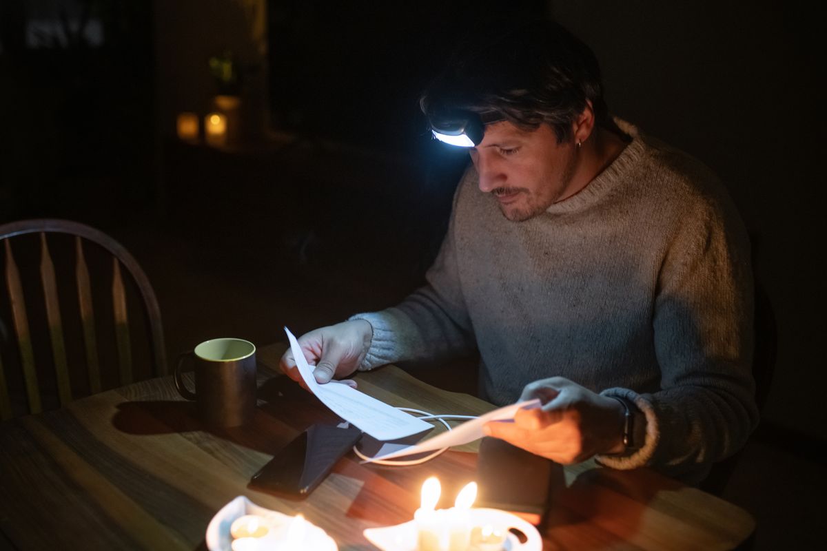 Man läser räkningar vid köksbordet i mörker, upplyst av pannlampa och ljus