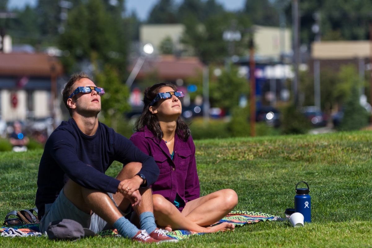 Två personer sitter på en filt i gräset och tittar mot himlen med skyddsglasögon