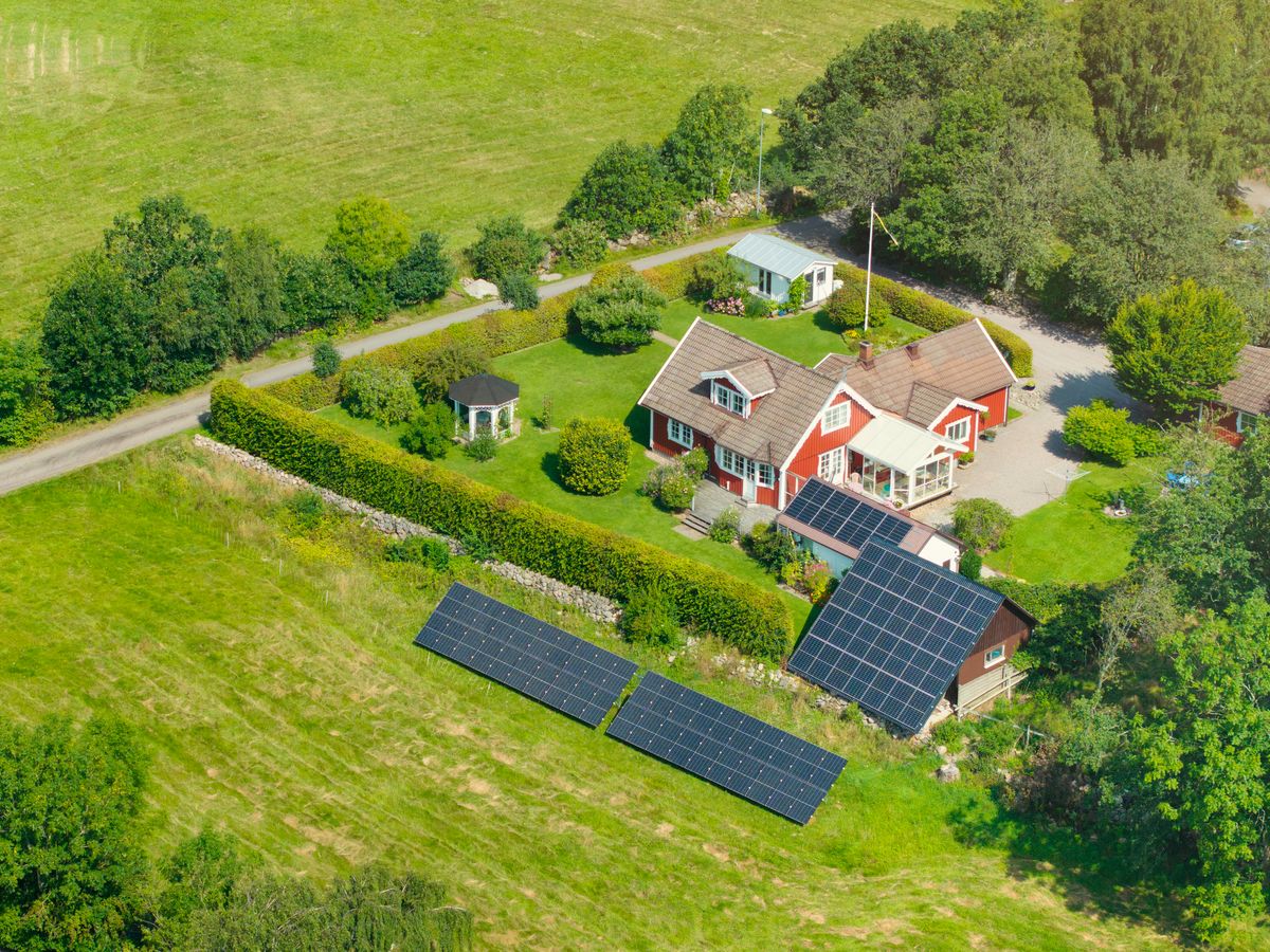 Solceller på tak och på mark. Rött hus på landet med solpaneler i trädgården omgivet av gröna åkrar