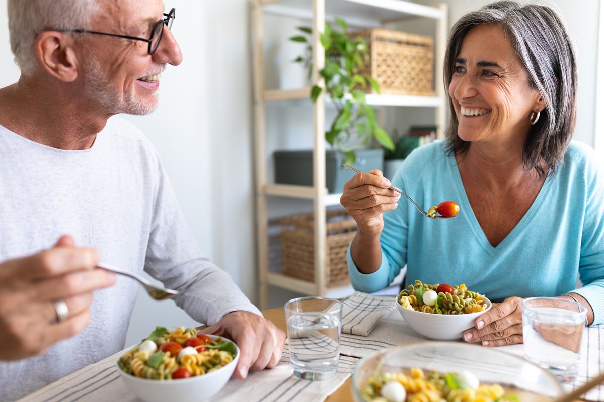 Äldre par sitter vid bord och äter sallad tillsammans, leende mot varandra
