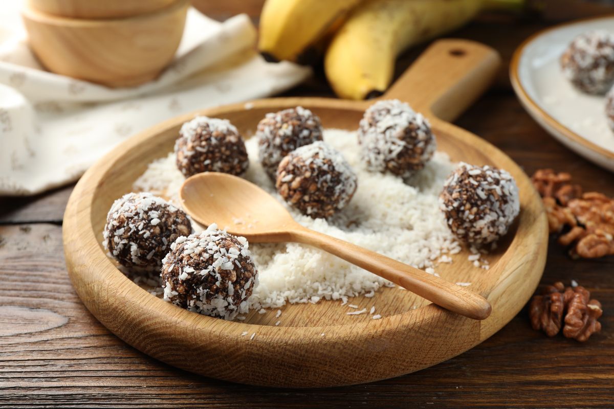 Chokladbollar rullade i kokos på träbricka med bananer och nötter i bakgrunden