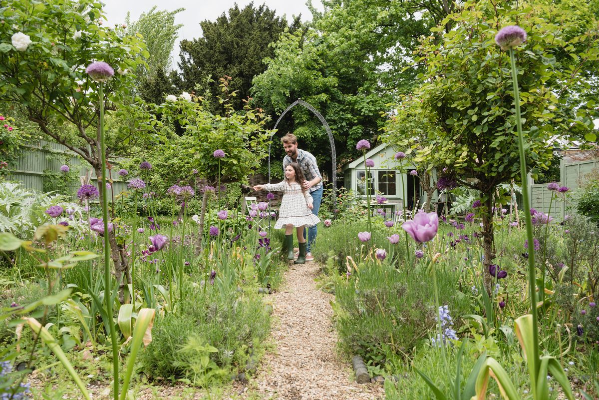 Pappa och dotter promenerar på grusgång genom en grönskande trädgård med blommor