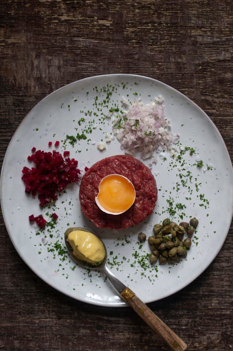 Råbiff på tallrik flankerad av en sked med senap, kapris, hackad lök och fint skuren rödbeta.