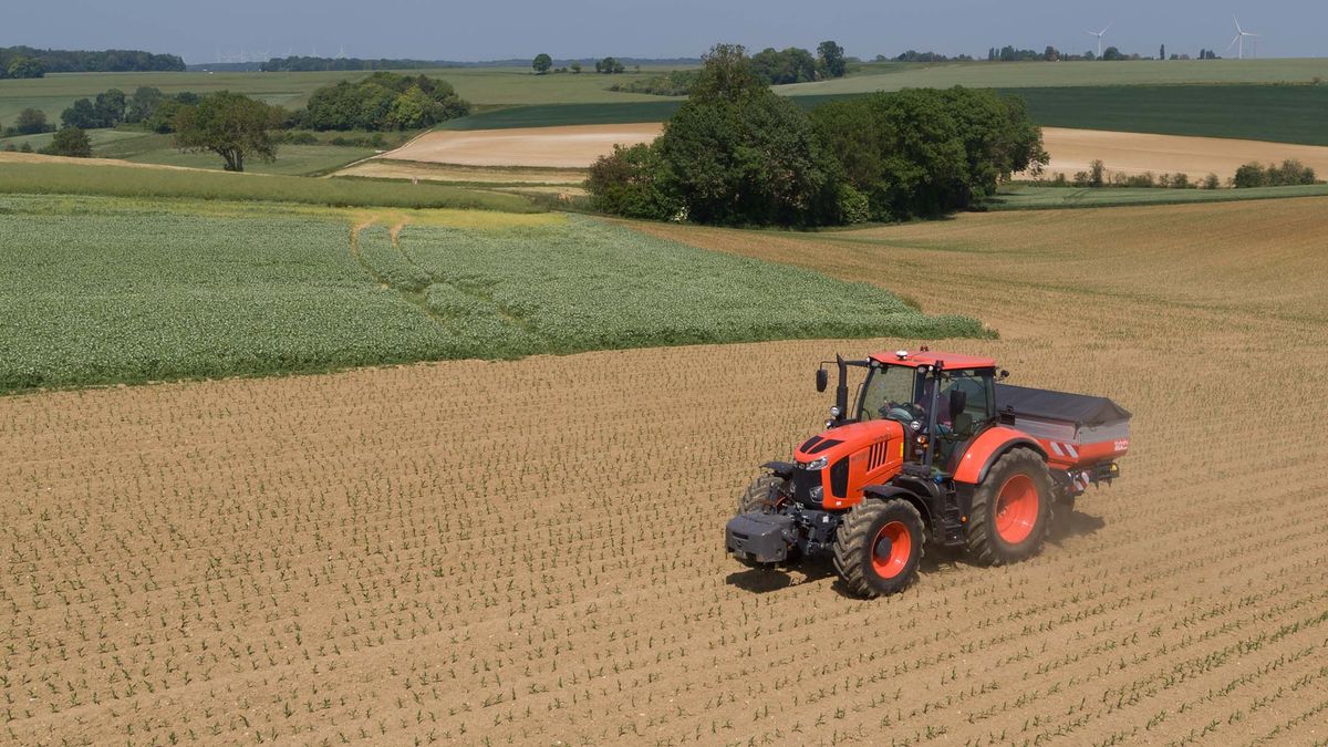 Orange traktor kör över åker med grödor i böljande jordbrukslandskap