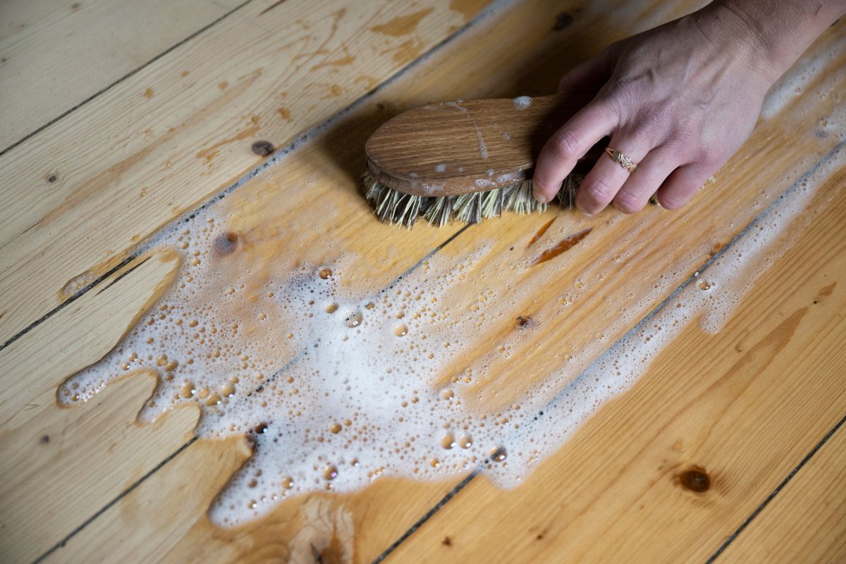Hand som skrubbar ett trägolv med skurborste och såpvatten