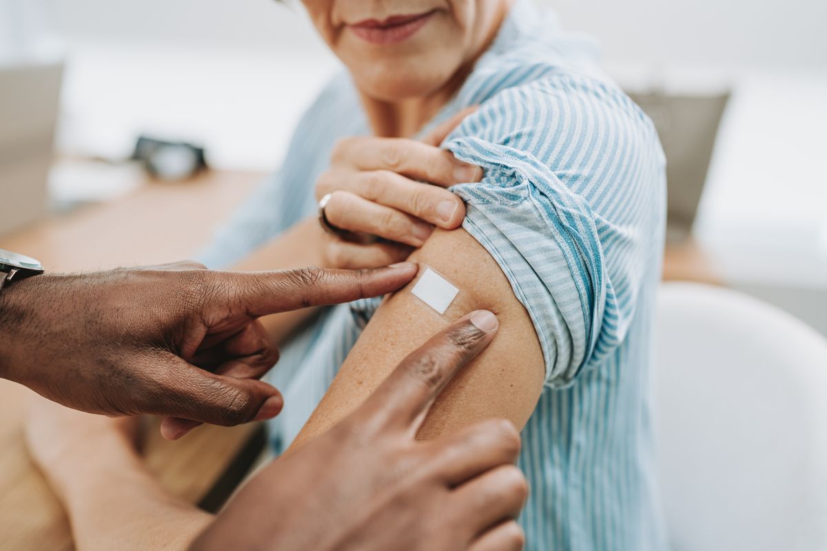 Kvinna sitter med upprullad ärm och plåster på armen efter vaccination. Ett par händer till man ur bild rör hennes arm.