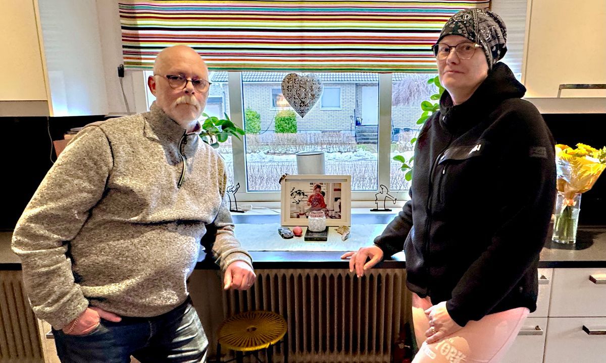 Två personer står i ett kök bredvid ett inramat foto och en bukett blommor